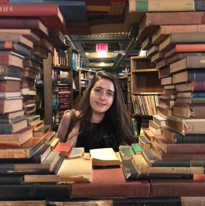 Kendra Kendra posing in a library with a circle of books stacked around her.